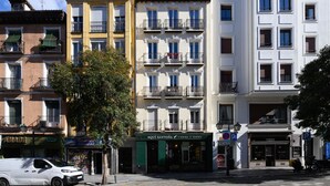 Un immeuble à plusieurs étages doté de balcons, un magasin avec une enseigne indiquant « AQUI SANTONA TIENDA Y TAPAS », et un fourgon blanc garé.