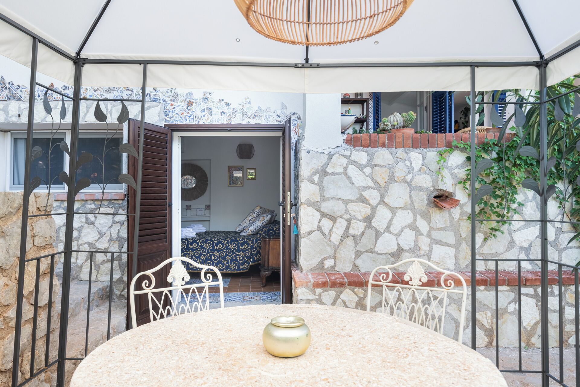 Een overdekt terras met een tafel en stoelen, een stenen muur en uitzicht op een kamer met een bed en een raam.