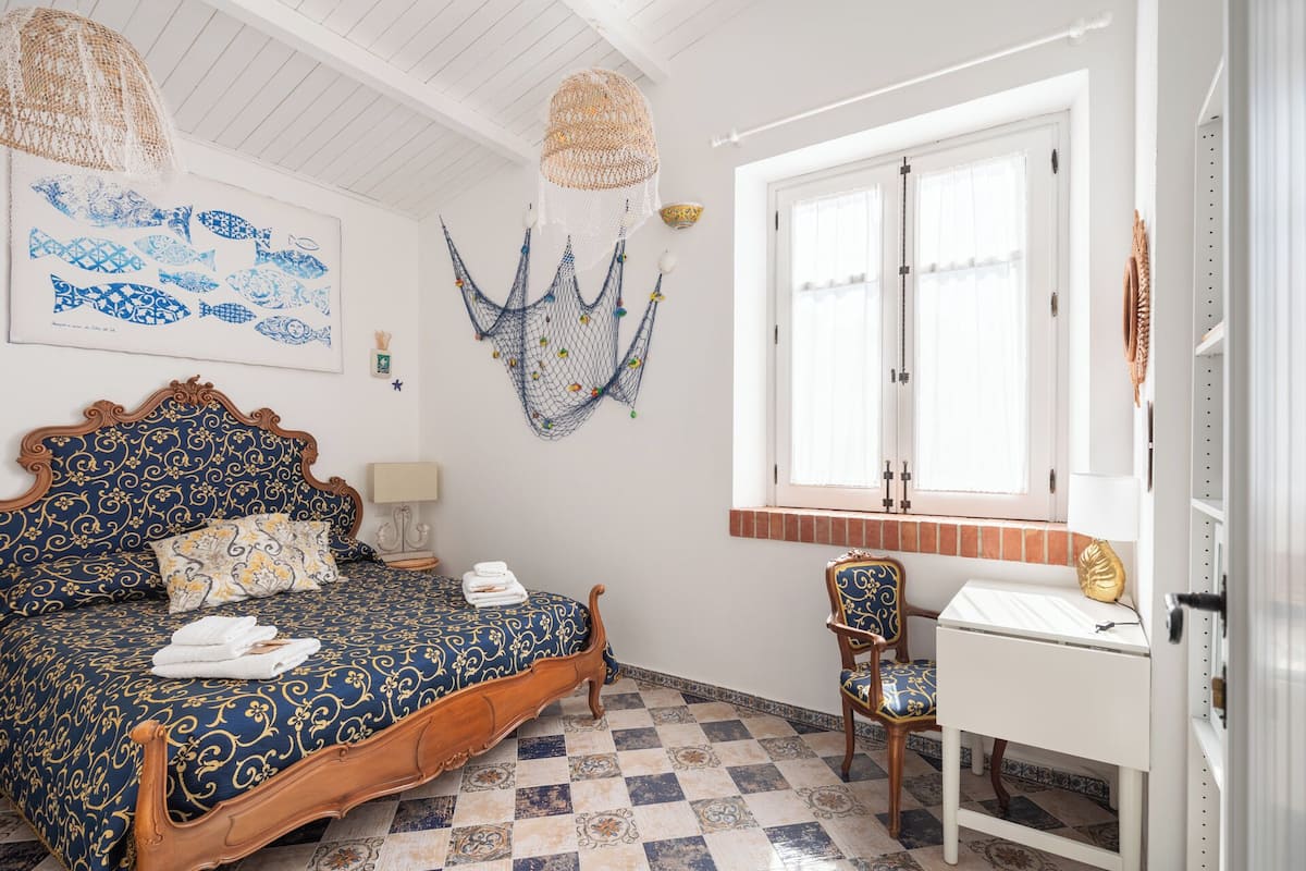 Ein Schlafzimmer mit kariertem Boden, einem Bett mit gemustertem Überwurf, einem Schreibtisch mit Stuhl und einem Fenster mit Jalousien.