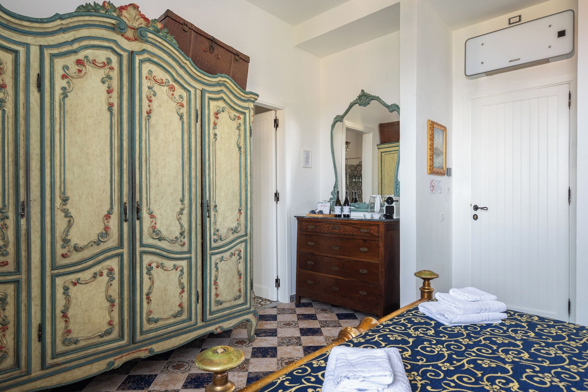 Una habitación con un armario grande y decorado, un aparador de madera, una cama con colcha estampada y un espejo en la pared.