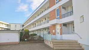 A multi-story residential building with a flat roof, a staircase leading to the entrance, and a small garden area with a hedge. - Relaxing vacation in the Panorama Whg. 8 - near the beach and the sea! (Grömitz)