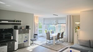 A modern living room with a grey sofa, a dining area with a table and chairs, a kitchen with a TV and cabinets, and large windows with curtains. - Relaxing vacation in the Panorama Whg. 8 - near the beach and the sea! (Grömitz)