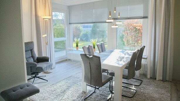 A modern dining room with a white table and grey chairs, large windows with blinds, and a view of greenery outside. - Relaxing vacation in the Panorama Whg. 8 - near the beach and the sea! (Grömitz)