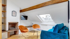 Ein modernes Loft mit freiliegenden Holzbalken, einem Kamin aus Backstein, orangefarbenen Sesseln, einem blauen Sofa und einem Dachfenster.