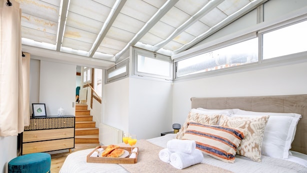 Ein Schlafzimmer mit einem großen Bett, einer Holzkommode, einem blauen Hocker, einem Frühstücksbrett und einem Fenster mit Blick nach draußen.