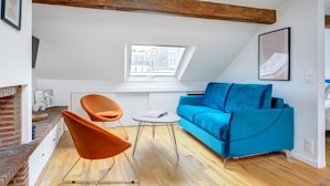 Ein modernes Loft mit freiliegenden Holzbalken, einem blauen Sofa, einem orangefarbenen Sessel und einem kleinen runden Tisch.