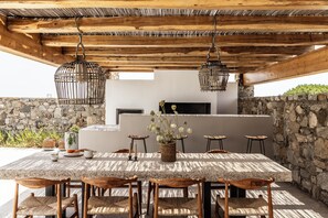 Una cocina al aire libre con pared de piedra, techo de madera y una mesa grande con sillas.