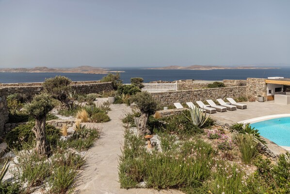 Un área de piscina con un sendero de piedra que lleva a sillones de descanso, rodeado de vegetación y vistas a un cuerpo de agua.
