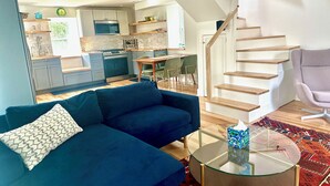 A modern living room with a blue sofa, a glass coffee table, and a staircase leading to an upper floor. - Charming 1-bedroom cottage on Wissahickon Park in Chestnut Hill (Philadelphia)