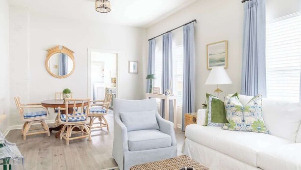Een woonkamer met een witte bank, een grijze fauteuil, een eettafel met rieten stoelen en een klein bijzettafeltje met een lamp.