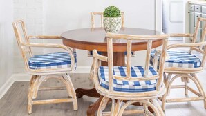 Een eetgedeelte met een ronde houten tafel, vier rieten stoelen met blauw-witte geruite kussens, een ronde spiegel met houten lijst, en een kleine potplant op de tafel.