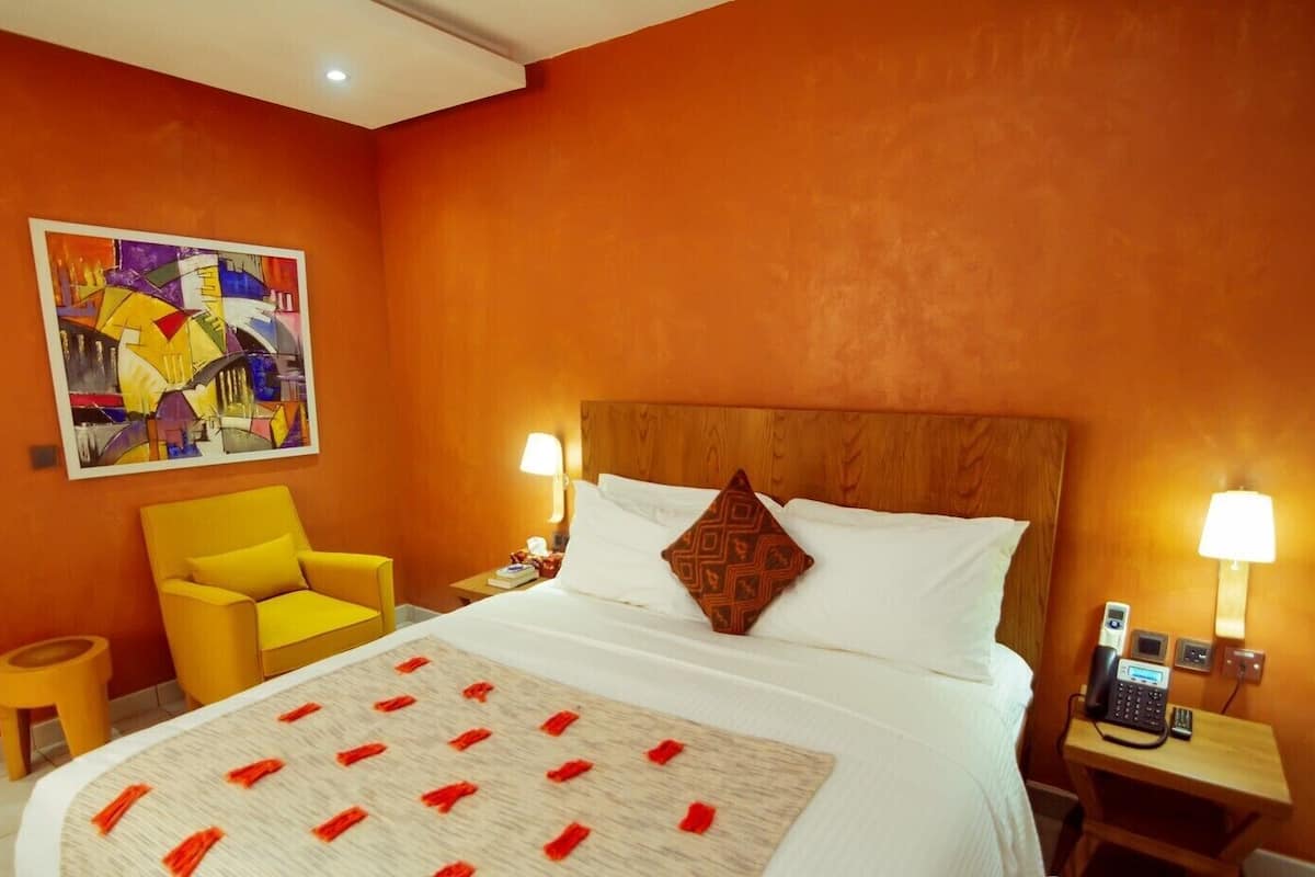 A hotel room with a large bed, a yellow armchair, a painting on the wall, and a nightstand with a phone and lamp.