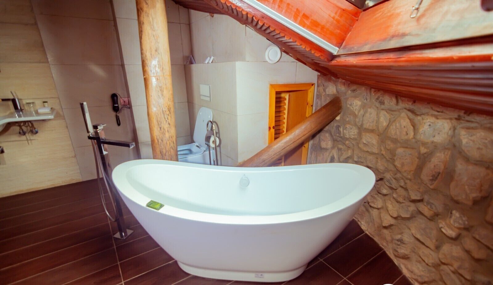 A modern bathroom with a freestanding bathtub, a wooden column, and a stone wall.