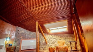 A cozy room with a sloped ceiling, a stone wall, a wooden chair, a small table with a lamp, and a window. - Trois Ore Residences  (Ibadan)