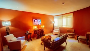 A room with orange walls, a TV, two lamps, a sofa, a chair, and a window with curtains. - Trois Ore Residences  (Ibadan)