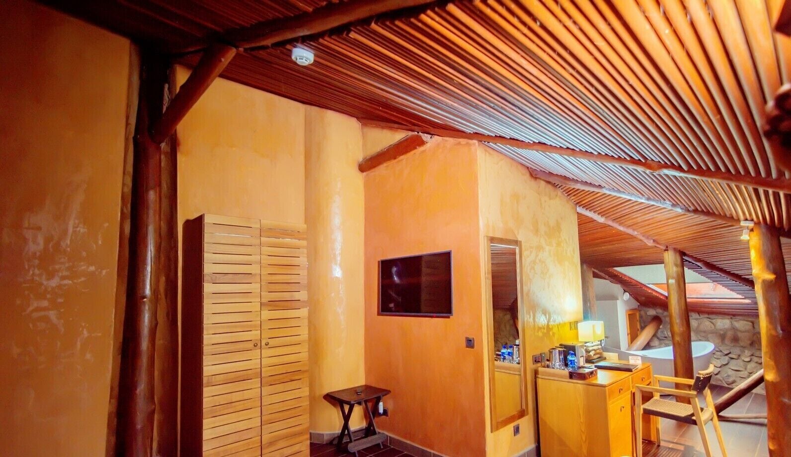 A room with wooden slatted ceilings, a wooden wardrobe, a small table with a lamp, and a mirror.