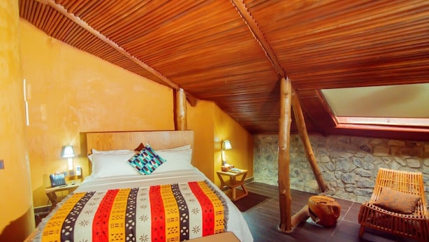 A cozy room with a sloped ceiling, a bed with a colorful blanket, a stone wall, and a wooden chair. - Trois Ore Residences  (Ibadan)