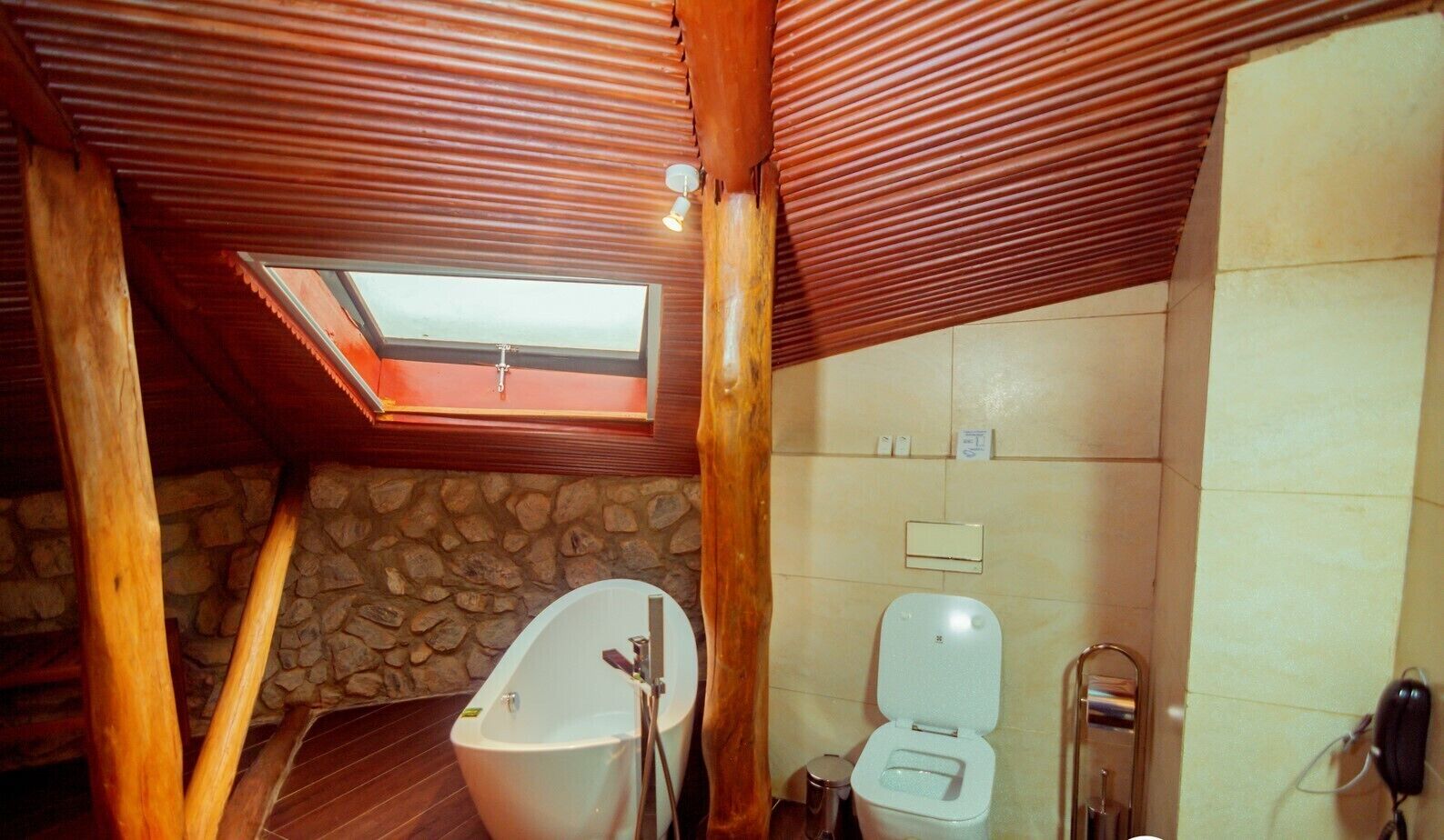 A bathroom with a stone wall, wooden beams, a skylight, a bathtub, and a toilet.