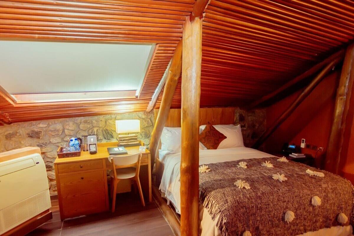 A cozy attic bedroom with a wooden ceiling, a desk with a computer, a bed with a floral bedspread, and a stone wall.