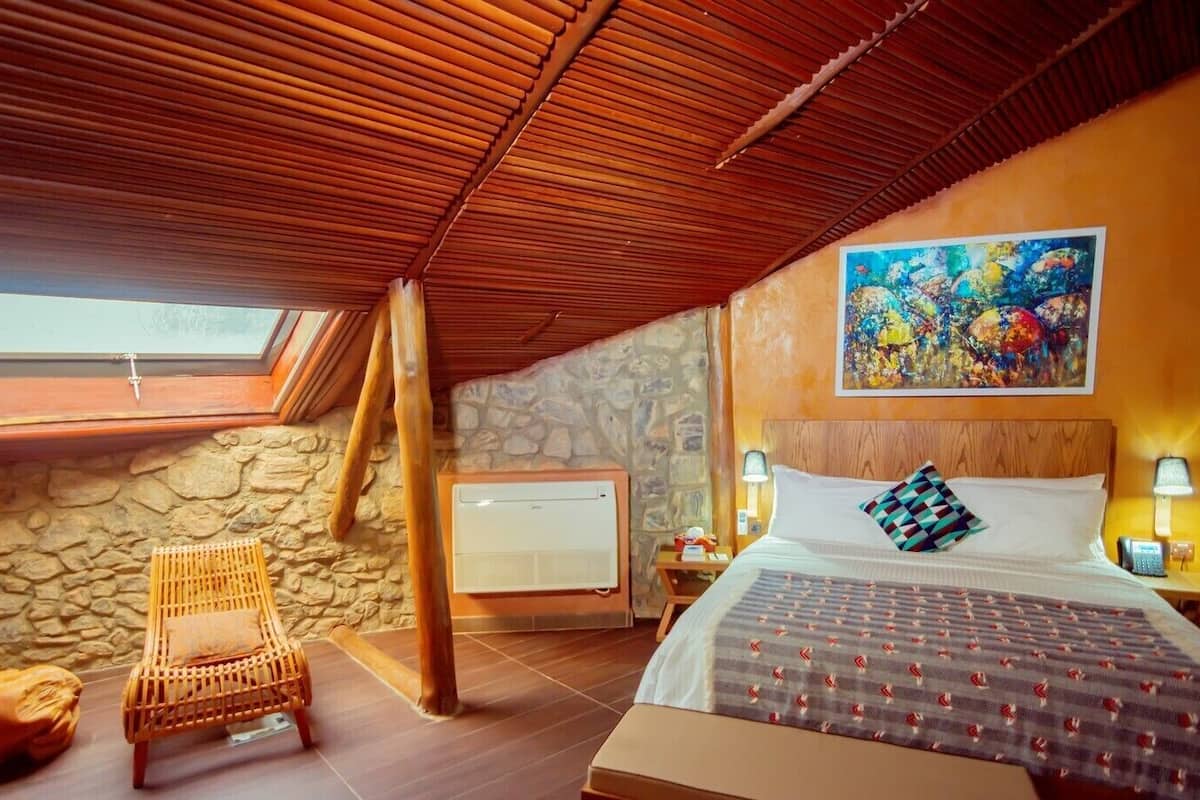 A cozy bedroom with a sloped ceiling, a stone wall, a wooden bed, a wicker chair, and a painting on the wall.
