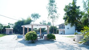 A covered outdoor seating area with a table and chairs, surrounded by small bushes and trees. - Badrika Farm's (Huzur)