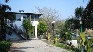 A building with a staircase leading to a covered area, surrounded by greenery and a body of water. - Badrika Farm's (Huzur)