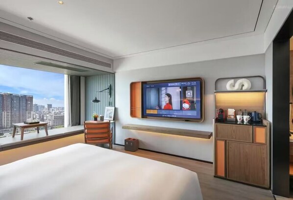 A modern hotel room with a large bed, a TV mounted on the wall, a small table with a chair, and a view of the city. - UrCove by Hyatt Shantou Xinyicheng Huanghe Road (Shantou)