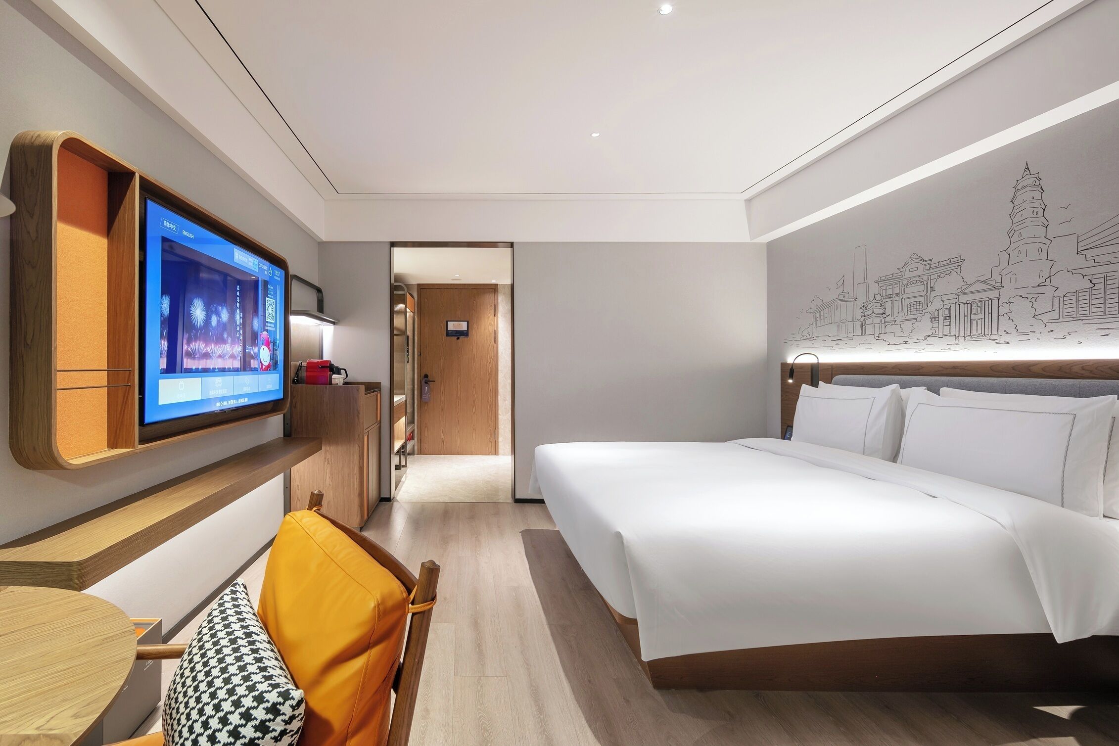 A hotel room with a bed, a TV mounted on the wall, a desk with a chair, and a hallway leading to another room.