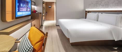 A hotel room with a bed, a TV mounted on the wall, a desk with a chair, and a hallway leading to another room.