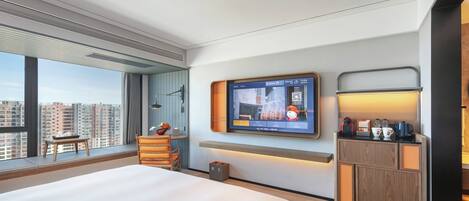 A modern hotel room with a large bed, a TV mounted on the wall, a small table with a chair, and a view of the cityscape.