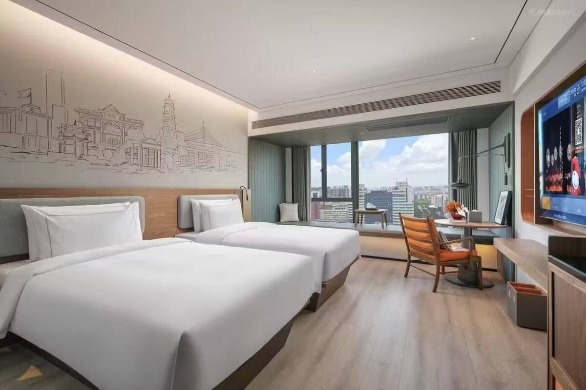 A hotel room with two beds, a desk, a chair, and a large window offering a city view.
