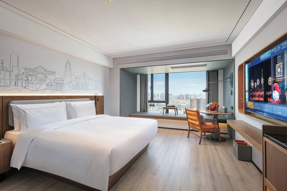 A modern hotel room with a large bed, a desk, a chair, a TV, and a city view.