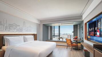 A modern hotel room with a large bed, a desk, a chair, a TV, and a city view.