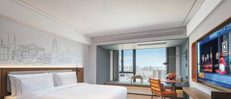 A modern hotel room with a large bed, a desk, a chair, a TV, and a city view.