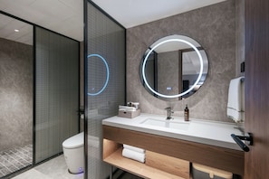 Un baño moderno con cabina de ducha de vidrio, un espejo redondo con iluminación LED, un lavamanos blanco y un mueble de tocador de madera.