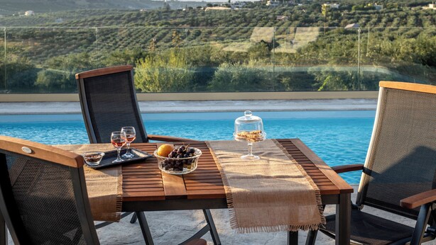 A dining table with chairs, a glass bowl of fruit, a wine glass, and a dessert on a terrace overlooking a pool and a scenic hillside. - Villa Plyto Amazing views Heated pool (Hersonissos)
