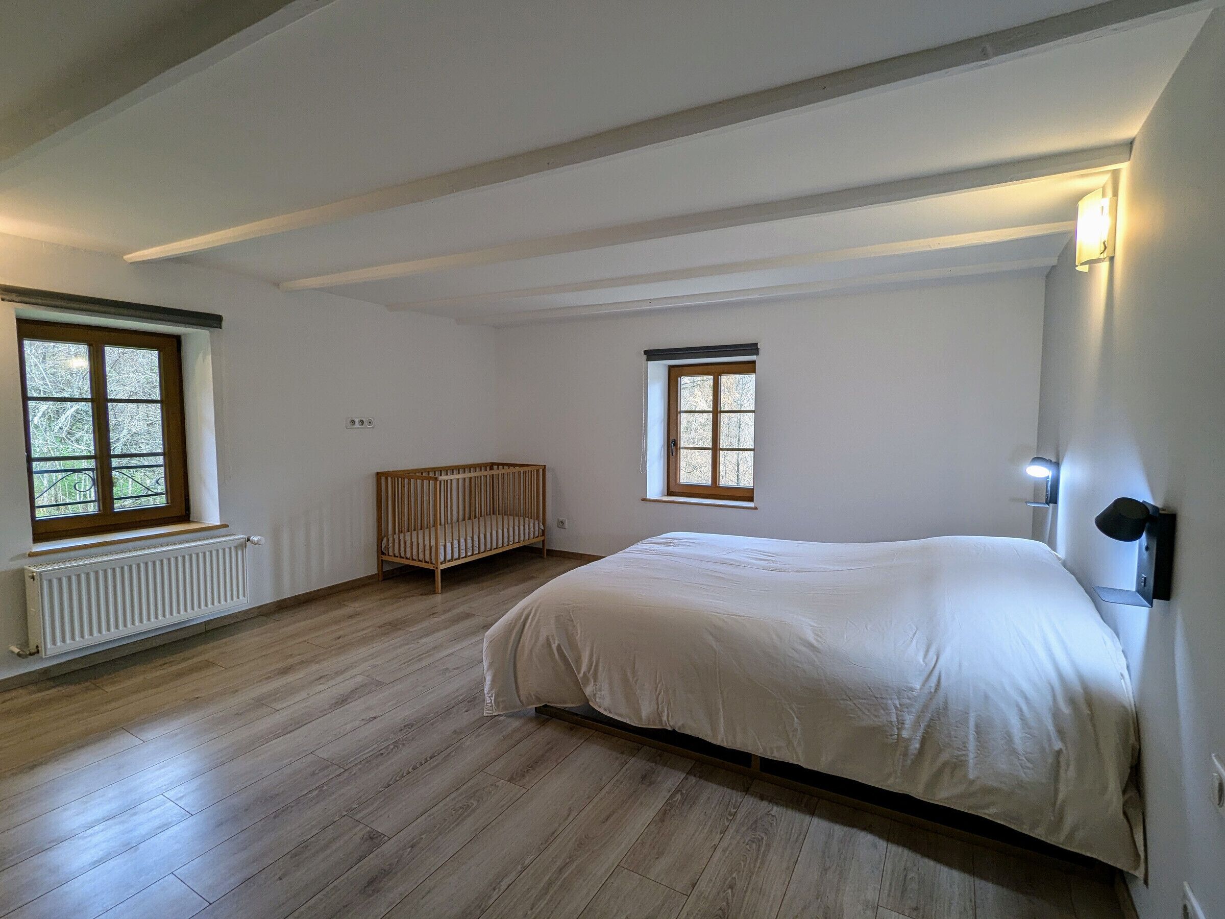 Un dormitorio con una cama grande, una cuna, dos ventanas y un radiador.