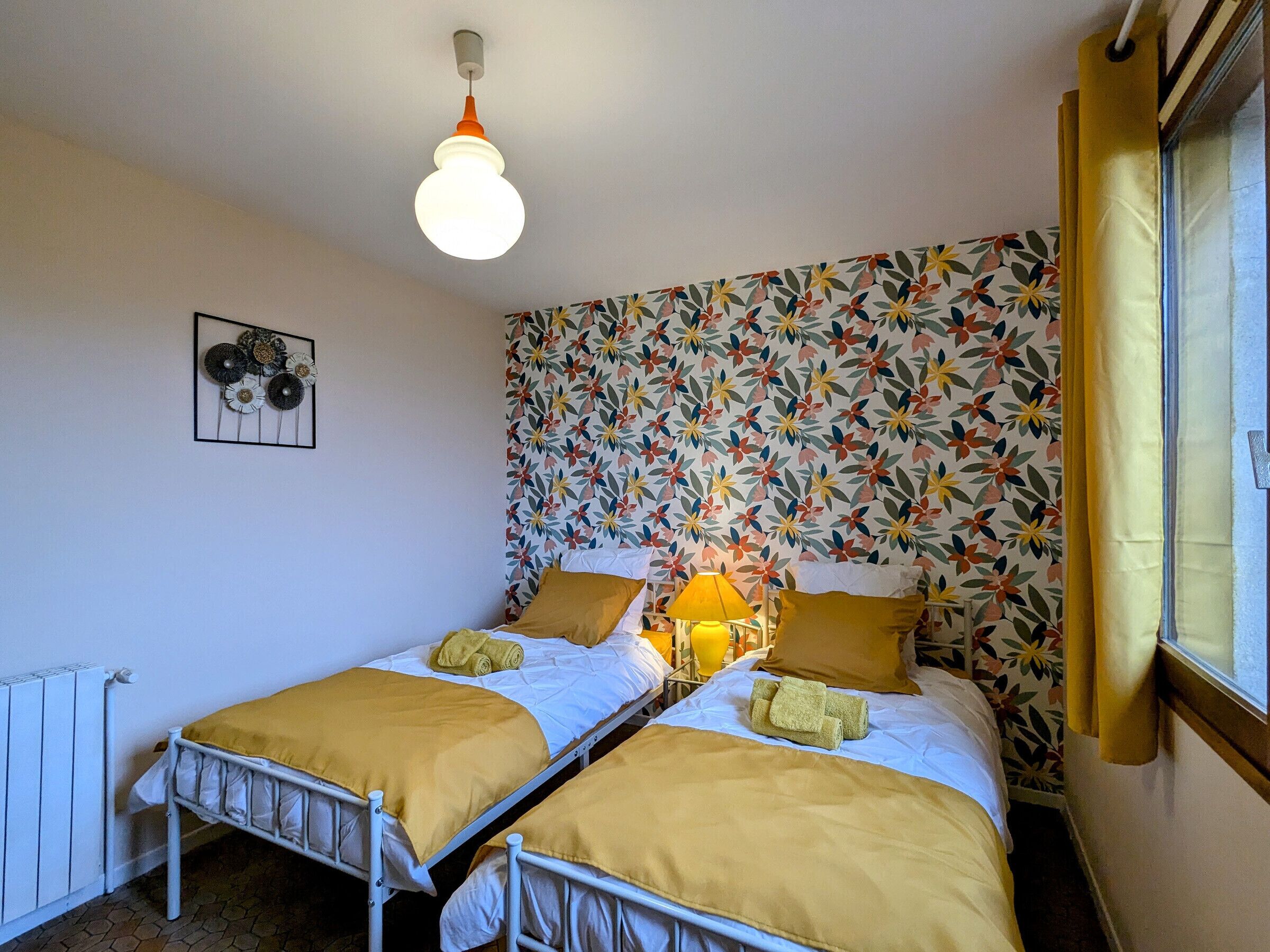 A room with two beds, a radiator, a wall-mounted light fixture, and a floral-patterned wallpaper.