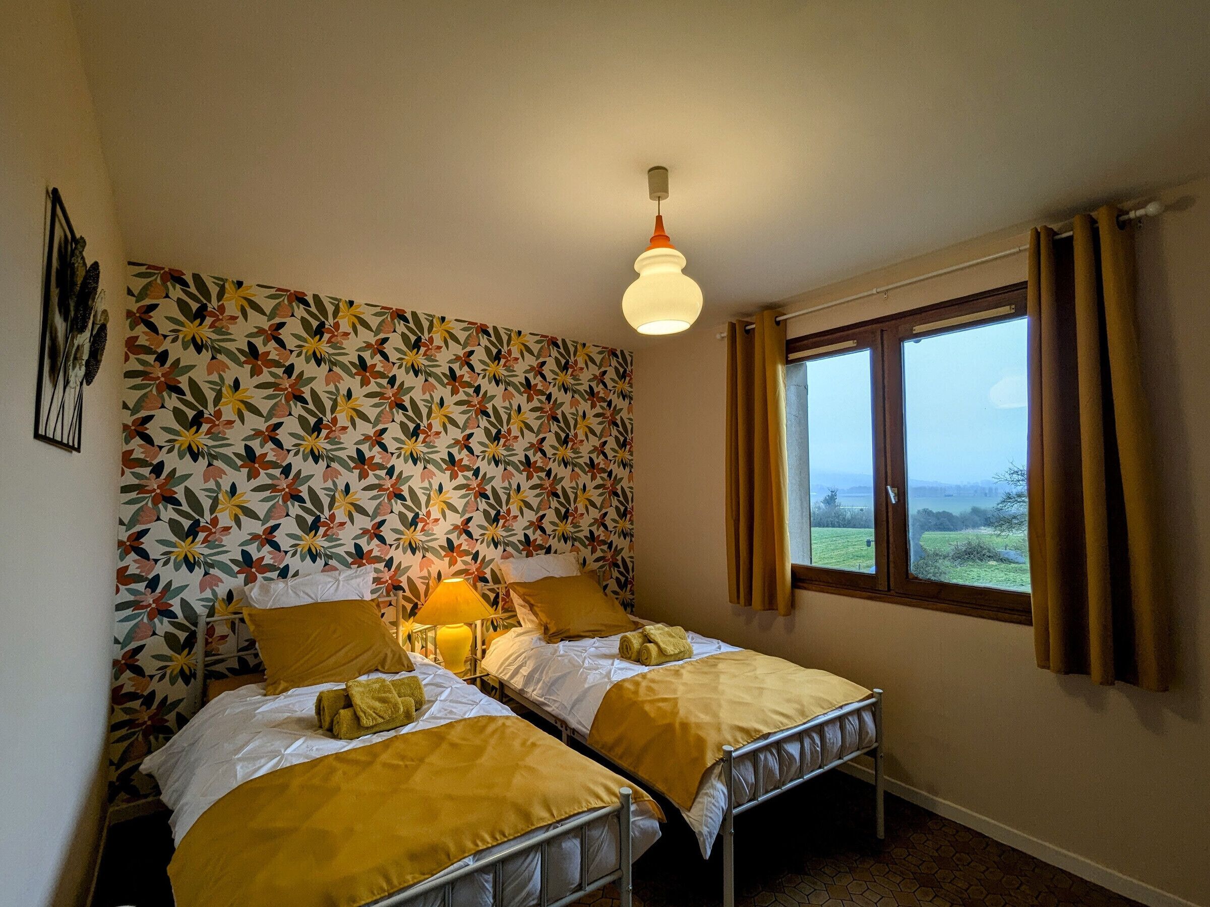 A room with two beds, a floral wallpaper, a window with curtains, and a hanging light fixture.