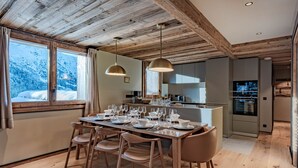 Un comedor con una mesa de madera dispuesta para seis personas, una cocina con electrodomésticos modernos y vistas a montañas nevadas a través de amplios ventanales.