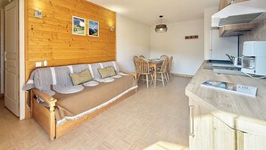 A cozy living area with a wooden wall, a sofa with striped cushions, a dining table with chairs, and a kitchen area with a coffee machine.