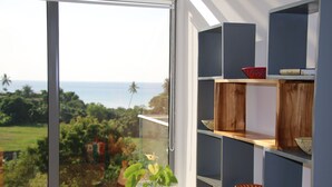 A room with a large window overlooking a scenic view of the ocean and greenery. There are wooden shelves with decorative items and a potted plant on the windowsill.