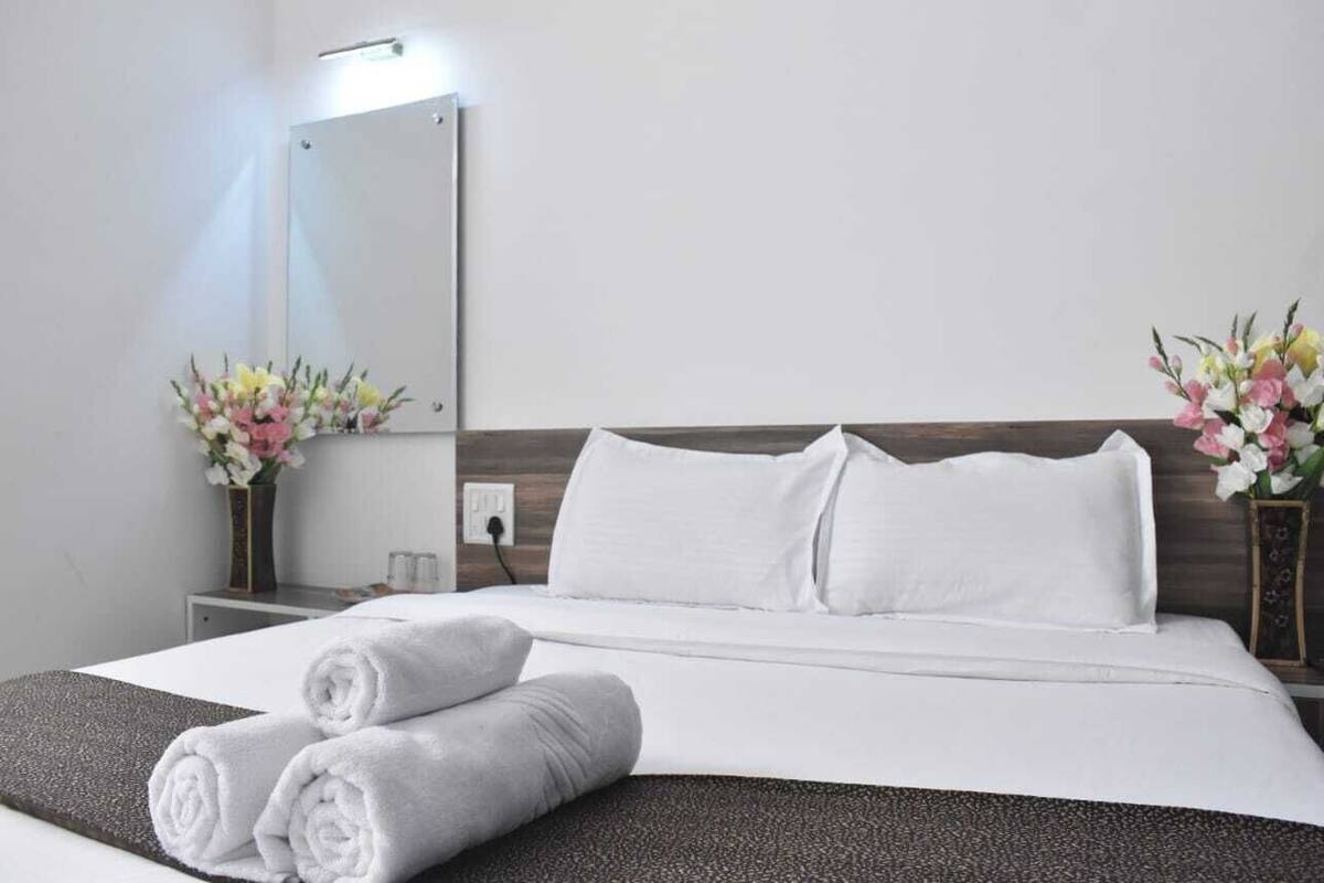 Una habitación de hotel con una cama bien hecha, almohadas blancas y un cabecero oscuro. Hay dos jarrones con flores a ambos lados de la cama.