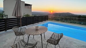 Un área de piscina en la azotea con una mesa y sillas, una sombrilla y vistas al atardecer sobre las colinas.