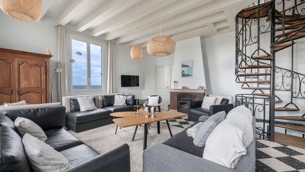 Een ruime woonkamer met leren banken, een houten salontafel en een zwart-wit geruit vloertje. Er is een open haard, een televisie en een trap met een smeedijzeren leuning.