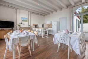 A modern kitchen with white cabinetry, a built-in oven, and a large island with seating. There is a dining area with white tables and chairs, and a large window providing natural light.