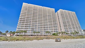 Un hotel moderno de gran altura con múltiples balcones, situado cerca de una playa con palmeras.