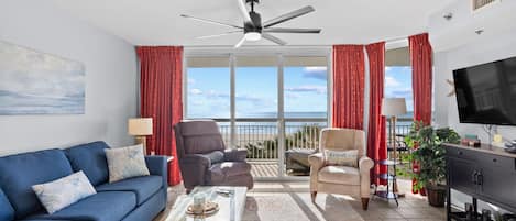 A living room with a blue sofa, a glass coffee table, a ceiling fan, a TV, and a view of the beach through large windows.