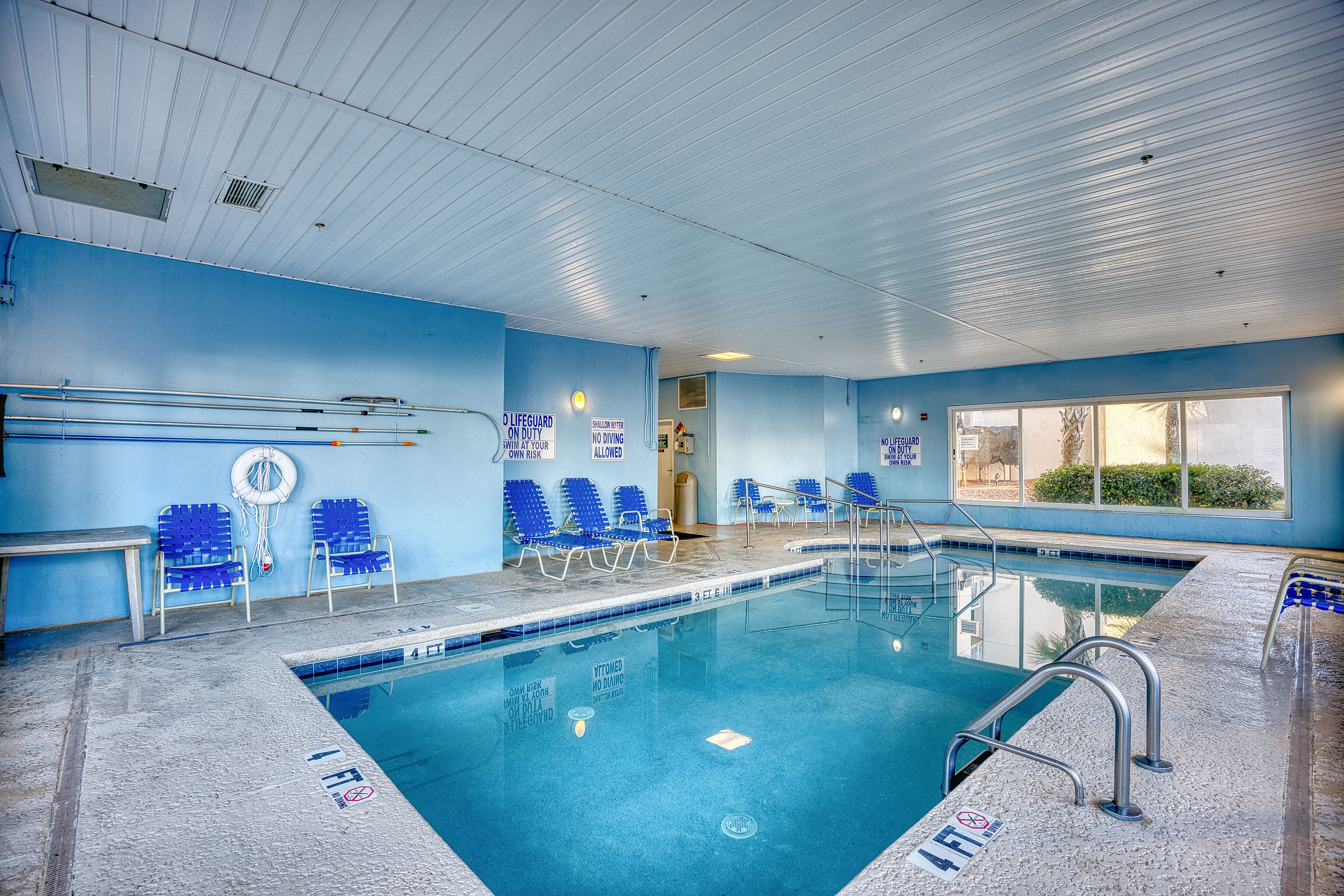 Une piscine aux murs bleus et au sol carrelé, entourée de chaises bleues et d’une plateforme pour les sauveteurs.