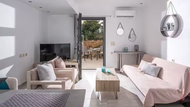 Un salon moderne avec un canapé, une table basse et un téléviseur. On aperçoit un coin repas avec une table et des chaises à travers une porte ouverte.